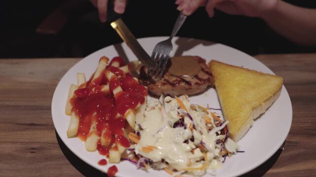 A steak being trimmed of its burnt parts with gravy sauce served with French fries and ketchup, creamy coleslaw salad, and toasted buttered bread on a white plate over