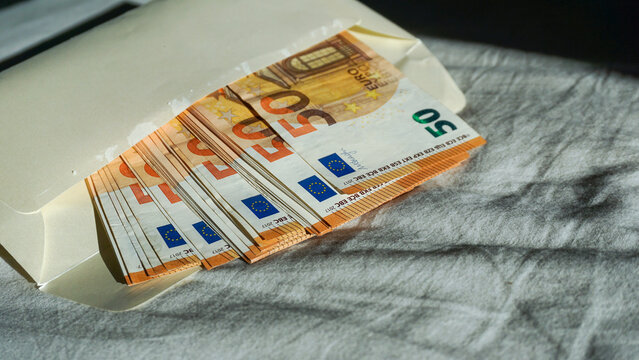 Stack of 50 euro banknotes partially hidden in envelope under dramatic shadow, suggesting cash payment, hidden money, shadow economy, tax evasion, corruption, bribery and illegal financial activity.