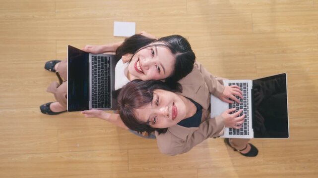 Top angle view of two happy Asian female colleagues sitting on the office floor with laptops, looking up at the camera and smiling broadly.