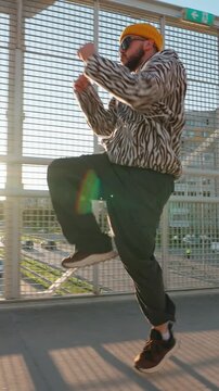 Close up young man in tiger print hoodie and yellow beanie dancing hip hop on street bridge with city background during sunset golden hour creative slow motion vertical video