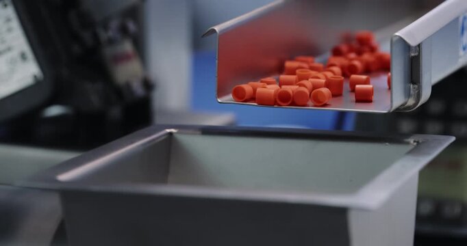 Automated feeder releases orange pharmaceutical capsules into collection bin. Controlled chute transfers uniform medicine units during factory processing
