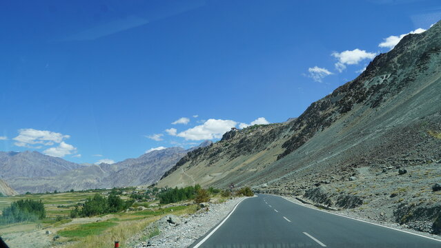 Road through mountainous landscape of zanskar, Ladakh, India with clear blue sky, green fields, rocky slopes and distant village under bright sunlight in summer season