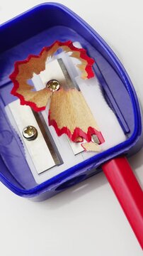 Pencil tip sharpening. Pencil sharpening inside blue sharpener. Red pencil wood curls expand around blade opening, showing steady use of small desk tool.