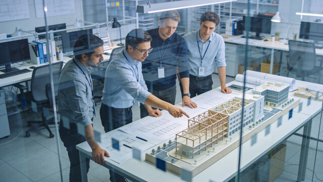 Professional architects collaborating on a modern building model inside a glass wall office environment with blueprints and technical documentation