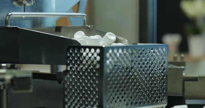 Lowering conveyor releases white medicine containers into collection area. Automated pharmaceutical line transfers empty bottles toward next packaging stage