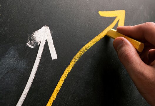 Hand draws upward trending arrows on a dark surface with yellow chalk