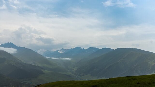 Static timelapse of green mountain valley in Kyrgyzstan. Low clouds drift as sky clears over distant peaks.