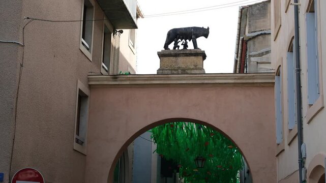 Narbonne, Occitania, France. August 26, 2026. Capitoline Wolf with Romulus and Remus on an arch in the heart of the historical tourist district. The street is decorated with ribbons fluttering in wind