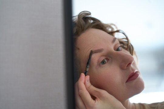 Woman grooming her eyebrows with a spoolie brush in front of a mirror.