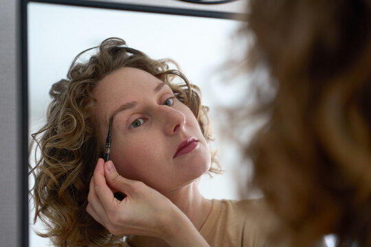 Woman grooming her eyebrows with a spoolie brush in front of a mirror.