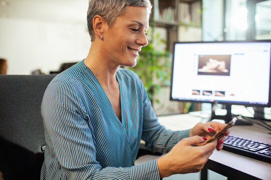 Smiling businesswoman using smartphone at computer desk in modern office