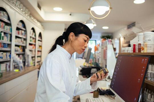 Pharmacist reviews medicine bottle at computer in modern pharmacy
