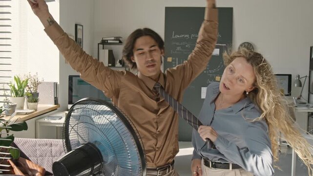 Asian young adult man and Caucasian young adult woman cooling down with electric fan at work. Suitable for HVAC service advertising, corporate summer campaigns and heatwave wellbeing content