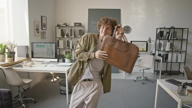 Joyful young adult Caucasian man dancing energetically with leather briefcase in modern open office. Suitable for business success, career achievement, or positive workplace culture campaigns