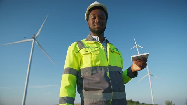 Serious African American male engineer wearing protective helmet and neon jacket standing outdoors holding miniature wind turbine. Observing wind farm and evaluating clean energy project in open field