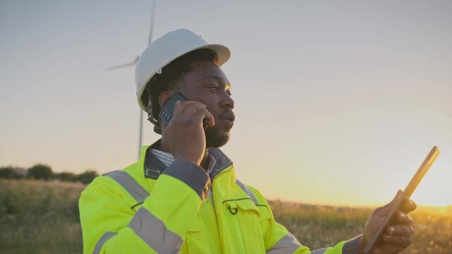African American male engineer in helmet and reflective jacket talking on smartphone while holding tablet outdoors. Standing near wind turbines during sunset and coordinating renewable energy tasks.