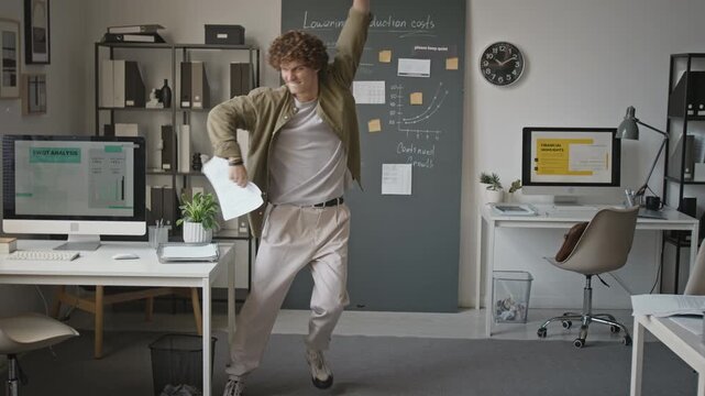 Caucasian young man dancing happily in office and throwing paperwork in air celebrating success. Ideal for corporate milestone campaigns, employee wellness promotions, business celebration videos