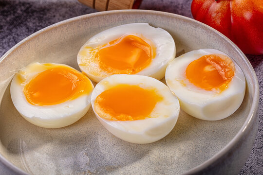 Four soft boiled egg halves with runny golden yolk in ceramic bowl, red tomato and wooden utensil in background, shallow depth of field