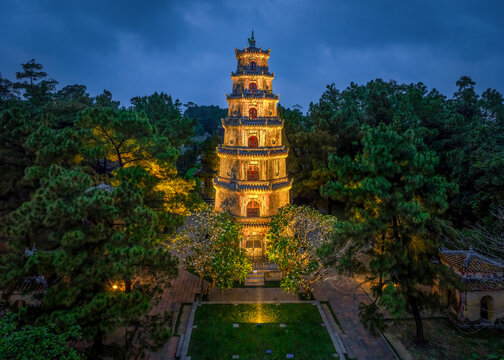 A majestic aerial night photograph of the historic Thien Mu Pagoda (Ch&ugrave;a Thi&ecirc;n Mụ) in Hue, Vietnam. The seven-story Phuoc Duyen Tower glows warmly against a deep blue dusk sky, framed by lush pine tre