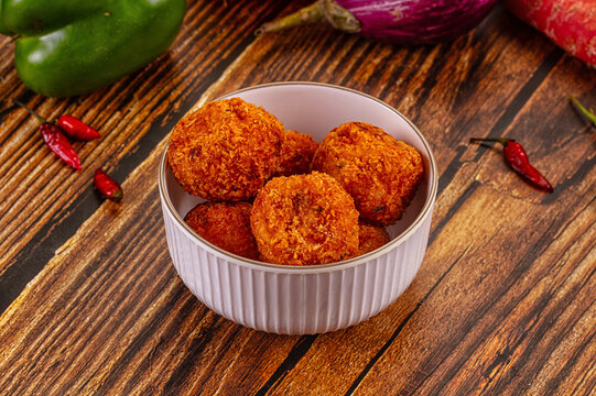 Crispy golden fried croquettes in white ceramic bowl on rustic wooden table with green pepper, red chili, and eggplant background
