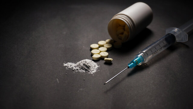 pills and syringe. Some pills spilled from the bottle, one syringe, and white powder on black background. Misused concept of drugs. 