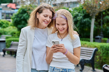 Happy mother and teenage daughter looking at smartphone together outdoors in city park