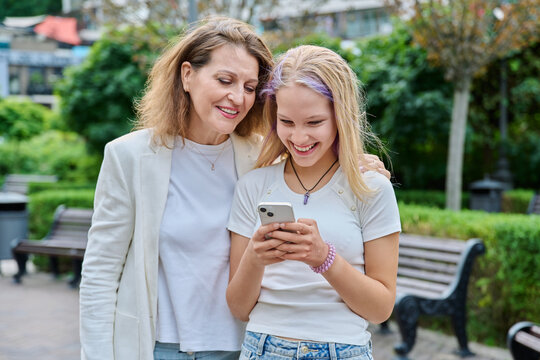 Happy mother and teenage daughter looking at smartphone together outdoors in city park
