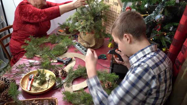 Grandmother and grandson make Christmas wreath together. Elderly woman and teenage boy decorate with their own hands. Family fun. DIY craft. Glue branches