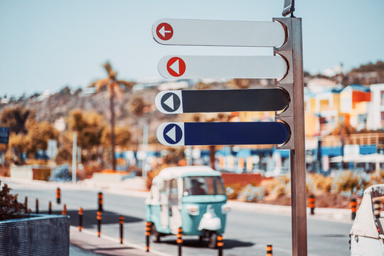Four empty wayfinding panels with left arrows offer templates for destinations or branding, sharply framed above a blurred turquoise tuk-tuk, palms and colorful buildings on a sunny street