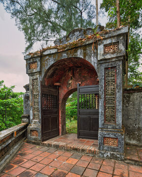 Weathered stone archway with intricate ornamental carvings and traditional wooden doors on a tiled terrace at the historical Thien Mu Pagoda in Hue, Vietnam