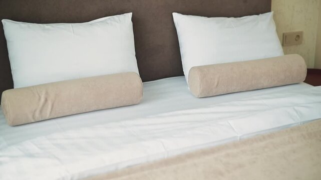 A bed is arranged with two white pillows placed against a brown wall. A rolled beige towel is positioned in front of the pillows on a plain white sheet