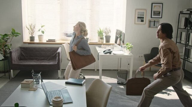 Caucasian woman and Asian man playfully sneaking and striking funny poses across office while finishing workday. Ideal for corporate culture promos and workplace wellness campaigns