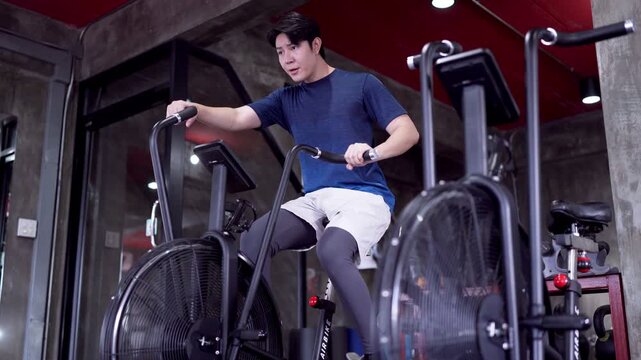 Young Asian man pumping air bike pedals during intense cardio workout in modern industrial gym