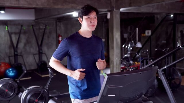 Young Asian man running on treadmill during workout session in industrial style gym at night