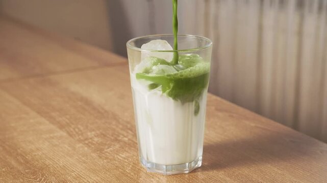 Pouring green matcha tea over iced milk in tall clear glass on wooden table. Preparing refreshing layered iced matcha green tea latte summer drink.
