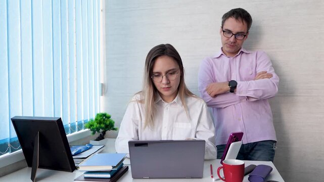 Boss or colleague supervising a female office worker typing on a laptop. Manager instructing subordinate, monitoring work, and giving directions.