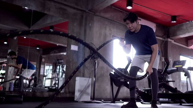 Young Asian man performing battle rope exercise in industrial style gym with dramatic lighting