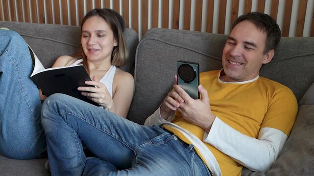 Bored couple on sofa at home: woman reading magazine, man using smartphone, each in their own world. Dull routine, lonely together, disengaged leisure.