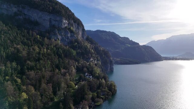 Aerial drone video of a Swiss alpine lake bordered by forested cliffs and rocky mountain slopes, with bright water reflections and distant valley views.