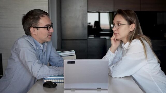 Man and woman at table with laptop and books, discussing work or study. Business meeting, home startup planning, or private tutoring.