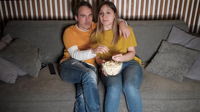 Young couple enjoying a romantic movie night on the sofa at home, watching an interesting film in the living room and sharing popcorn during a cozy date.
