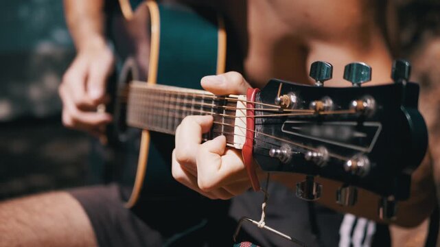 Close up of a musician fingerpicking acoustic guitar chords with a capo attached on the fretboard outdoors. Detailed focus showcases hand placement and string manipulation.