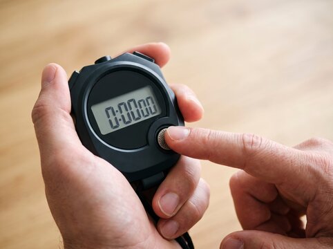Close-up of male hands holding a black digital electronic stopwatch, pressing the start button with a display showing zero on a wooden background.