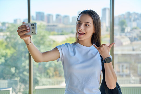 Smiling female student taking selfie photo showing shaka sign city window