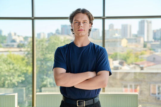 Confident young man standing with crossed arms looking at camera near big window