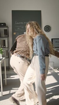 Vertical video of Asian man and Caucasian woman shaking hands and dancing playfully with folder in office. Footage fits corporate culture campaigns and employee satisfaction promotions