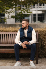 Obraz premium Taking a moment to relax outdoors, a casually dressed man sits comfortably in a stylish vest surrounded by autumn foliage. The cozy setting, natural light and warm colors create a calm seasonal atmosp