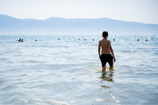 Boy walking into calm sea on sunny day, real lifestyle beach scene with mountains in background and natural light reflection.