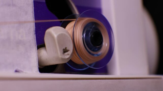 Sewing machine bobbin spinning rapidly during thread winding process. Detailed view shows clear plastic bobbin holder and beige thread being wound.
