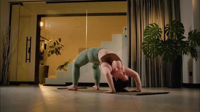 Woman holding bridge pose during yoga practice inside modern home with plants and warm evening glows